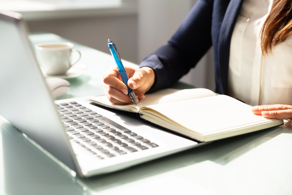 The image depicts a person, dressed professionally in a business suit, writing in a notebook with a pen. A laptop is placed in front of them on a glass-top desk, accompanied by a cup of coffee. The setting suggests a workspace, with natural light illuminating the scene.